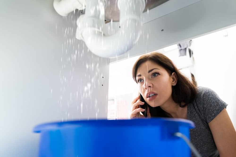 Emergency plumbers Pete the Plumber in Calgary Alberta available 24/7 portrayed by a women on the phone after a pipe broke, under the counter, filling a blue bucket.